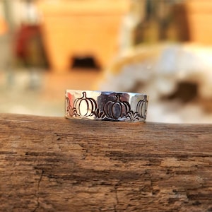 May include: A silver ring with a hammered finish and a design of pumpkins around the band.