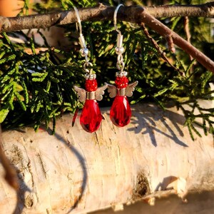 May include: Red glass angel wing earrings with silver accents. The earrings are hanging from a branch.