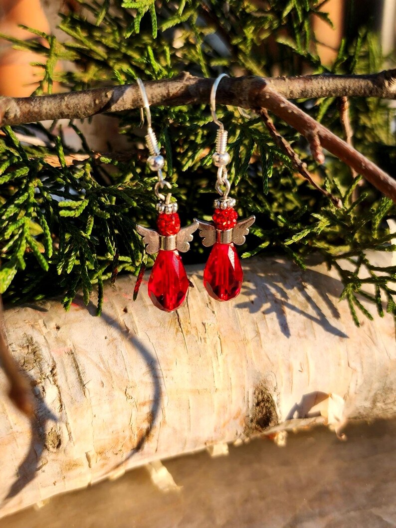 May include: Red glass angel wing earrings with silver accents. The earrings are hanging from a branch.