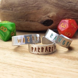 May include: Three silver rings with the words "WIZARD", "RANGER", and "BARBARIAN" stamped on them. A green and a red die are in the background. The rings are on a wooden surface.