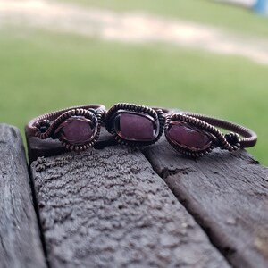 May include: Three copper wire wrapped rings with pink gemstone accents. The rings are arranged in a row on a wooden surface.