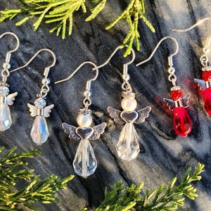May include: Five pairs of dangle earrings featuring angel charms. Two pairs are clear crystal with silver accents, one pair is clear crystal with a white pearl accent, and two pairs are red crystal with silver accents.
