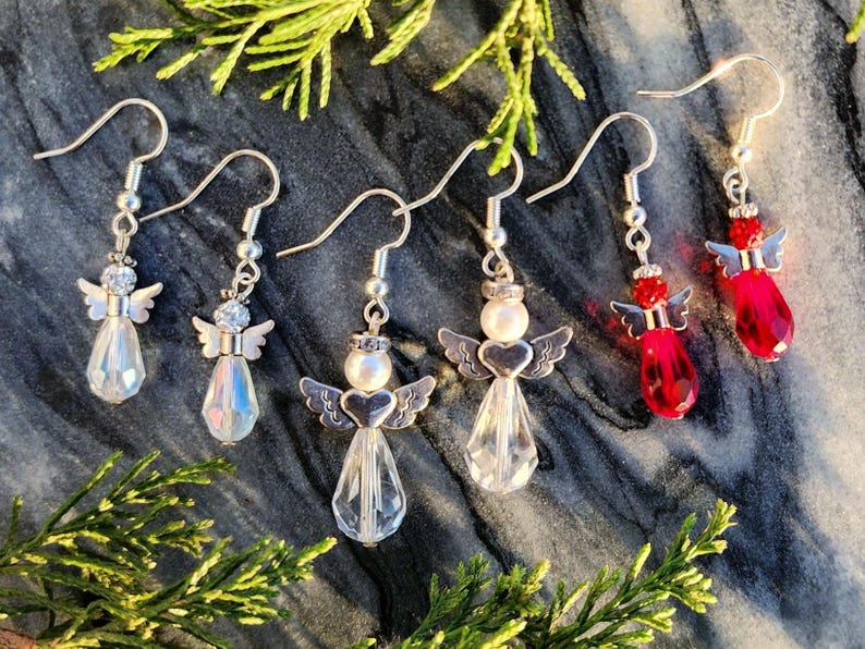 May include: Five pairs of dangle earrings featuring angel charms. Two pairs are clear crystal with silver accents, one pair is clear crystal with a white pearl accent, and two pairs are red crystal with silver accents.