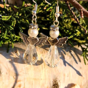 May include: Silver dangle earrings featuring a small angel with wings and a pearl bead. The angel is holding a heart and the earrings have a crystal teardrop pendant.