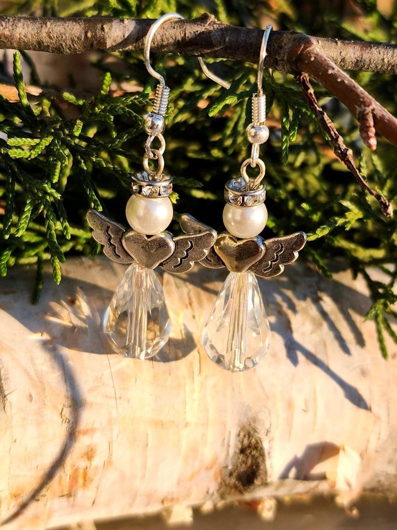 May include: Silver dangle earrings featuring a small angel with wings and a pearl bead. The angel is holding a heart and the earrings have a crystal teardrop pendant.