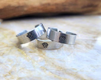 May include: Three silver-toned adjustable rings with engraved floral designs. One ring features a daisy, another a rose, and the third a small flower. The rings are displayed on a marble-like surface.