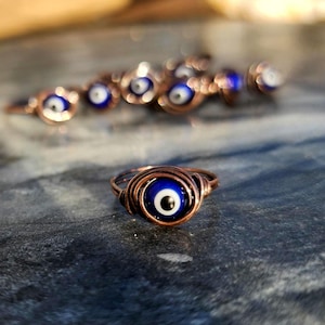 May include: A copper wire ring with a blue evil eye charm. The ring is wrapped in copper wire and the evil eye charm has a white center and a black pupil.