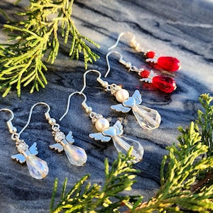 May include: Four pairs of dangle earrings featuring silver angel charms with crystal accents. Two pairs have white pearl accents and two pairs have red crystal accents.
