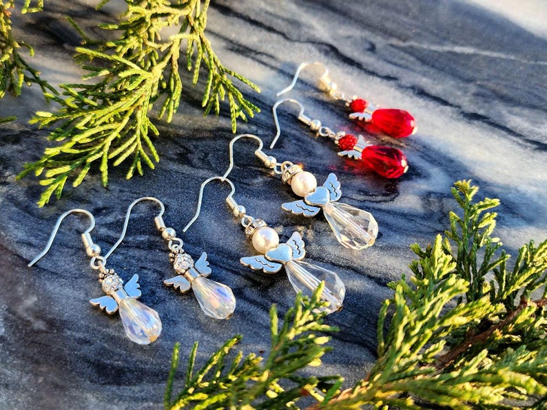 May include: Four pairs of dangle earrings featuring silver angel charms with crystal accents. Two pairs have white pearl accents and two pairs have red crystal accents.