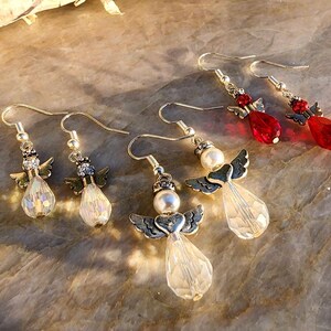 May include: Four pairs of angel-shaped dangle earrings. Two pairs feature clear crystal beads and silver angel wings. The other two pairs feature red crystal beads and silver angel wings.
