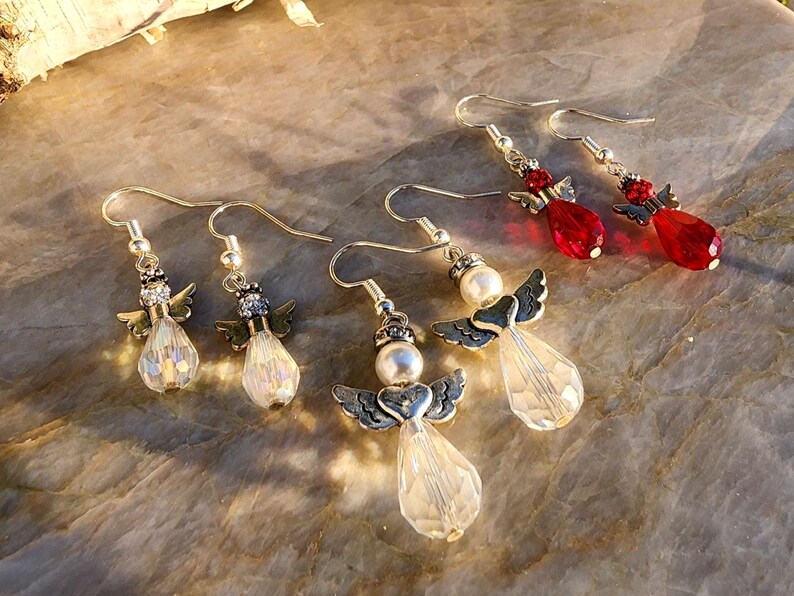 May include: Four pairs of angel-shaped dangle earrings. Two pairs feature clear crystal beads and silver angel wings. The other two pairs feature red crystal beads and silver angel wings.
