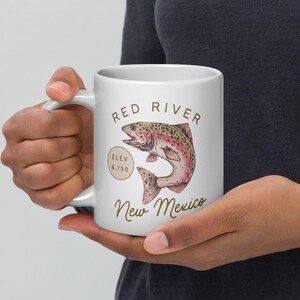 May include: White ceramic mug featuring a detailed illustration of a fish with the text "Red River New Mexico". The mug also includes the elevation of 8,750 feet. The design is in shades of brown, pink, and green.