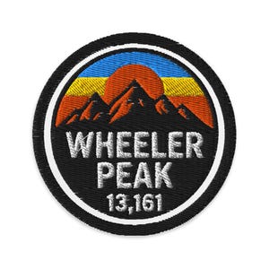 May include: Embroidered patch featuring a mountain range silhouette with a setting sun in shades of orange, yellow, and blue. The text "WHEELER PEAK" is in white, with "13,161" below. The patch has a black background with a white border.