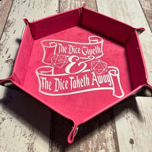 May include: A pink hexagonal dice tray with a white felt lining. The tray has a white inscription that reads "The Dice Giveth & The Dice Taketh Away" with two dice images.