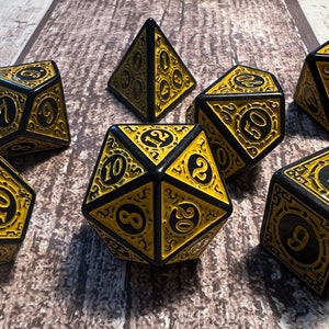 May include: A collection of seven black and gold polyhedral dice. Each die has gold numbering and decorative patterns. These dice are designed for role-playing games and are displayed on a wooden surface.