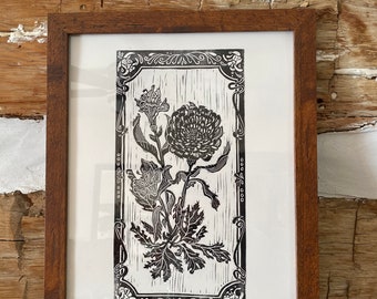 May include: A black and white woodblock print of a flower in a decorative frame. The flower has a large, round head and a stem with leaves. The frame is ornate and has a floral design.
