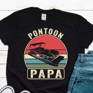 May include: Black t-shirt with a retro design featuring a pontoon boat illustration inside a circular graphic with the words "PONTOON PAPA". The shirt is paired with blue jeans and white sneakers.