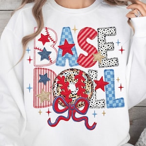 Puede incluir: Sudadera blanca con la palabra "BASEBALL" en un diseño de patchwork. Las letras son en rojo, blanco y azul con acentos de estrellas y béisbol. La letra "O" es una pelota de béisbol con un lazo rojo.