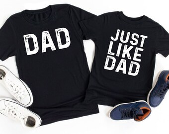 May include: Two black t-shirts with white text. The larger shirt says "DAD" and the smaller shirt says "JUST LIKE DAD".