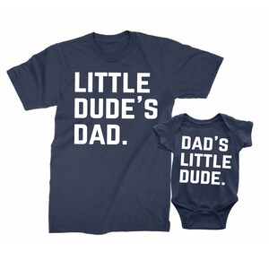 May include: Navy blue t-shirt with the text "LITTLE DUDE'S DAD." next to a navy blue baby onesie with the text "DAD'S LITTLE DUDE." Both items have white lettering. The items are likely a matching set.