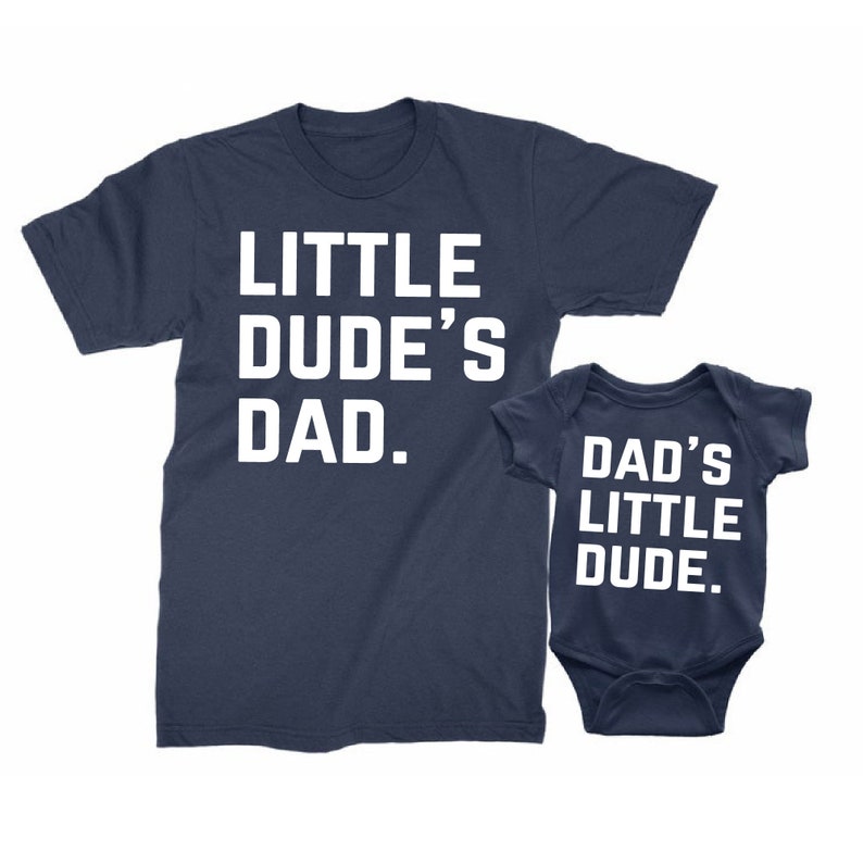 May include: Navy blue t-shirt with the text "LITTLE DUDE'S DAD." next to a navy blue baby onesie with the text "DAD'S LITTLE DUDE." Both items have white lettering. The items are likely a matching set.