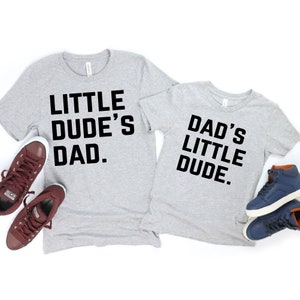 May include: Two light gray t-shirts with black text that reads "LITTLE DUDE'S DAD." and "DAD'S LITTLE DUDE." Maroon and navy blue sneakers are placed near the shirts. The shirts are a matching set.