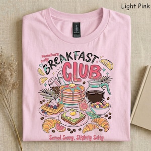 Pode incluir: Camiseta rosa claro com um gr&aacute;fico colorido de alimentos de caf&eacute; da manh&atilde;, incluindo panquecas, waffles e croissants. O texto "Breakfast Club" &eacute; exibido acima das ilustra&ccedil;&otilde;es de alimentos, com a frase "Served Sunny, Slightly Salty" abaixo.