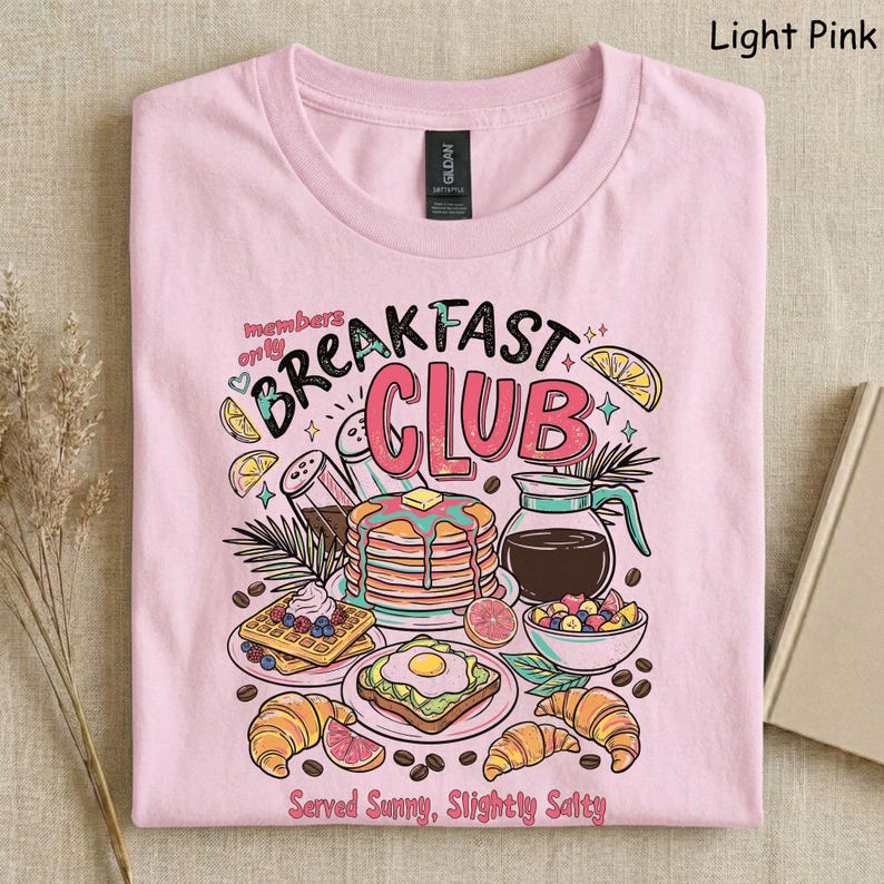 Pode incluir: Camiseta rosa claro com um gr&aacute;fico colorido de alimentos de caf&eacute; da manh&atilde;, incluindo panquecas, waffles e croissants. O texto "Breakfast Club" &eacute; exibido acima das ilustra&ccedil;&otilde;es de alimentos, com a frase "Served Sunny, Slightly Salty" abaixo.