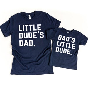 May include: Two navy blue t-shirts with white text. One shirt reads "LITTLE DUDE'S DAD." The other shirt reads "DAD'S LITTLE DUDE." Both shirts are short-sleeved and made of a soft material.