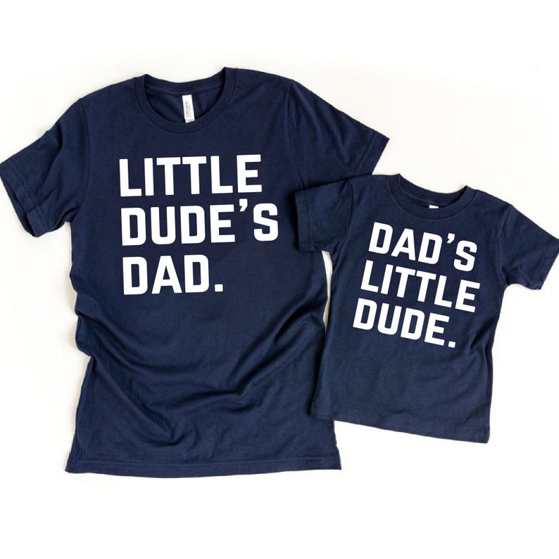 May include: Two navy blue t-shirts with white text. One shirt reads "LITTLE DUDE'S DAD." The other shirt reads "DAD'S LITTLE DUDE." Both shirts are short-sleeved and made of a soft material.
