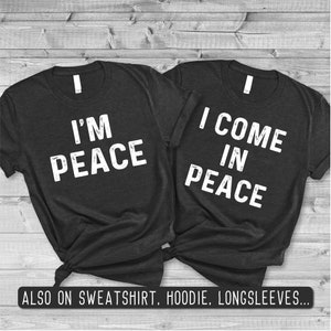 Può includere: Due magliette nere con testo bianco. Una maglietta dice "I'm Peace" e l'altra dice "I Come In Peace".