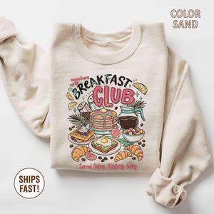 Pode incluir: Camisola cor de areia com um gr&aacute;fico colorido de alimentos de pequeno-almo&ccedil;o, incluindo panquecas, waffles e croissants. O texto "Breakfast Club" &eacute; exibido de forma proeminente, juntamente com a frase "Served Sunny, Slightly Salty."