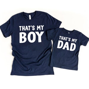 May include: Two navy blue t-shirts with white text. The larger shirt says "That's My Boy" and the smaller shirt says "That's My Dad".