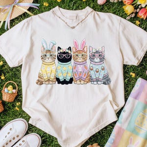 May include: A cream-coloured t-shirt featuring four cats dressed for Easter. Each cat wears glasses and a sweater with Easter-themed designs. The cats have bunny ears or floral crowns. The shirt is on a grassy surface with Easter decorations.
