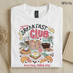 Pode incluir: Camiseta branca com um gr&aacute;fico colorido de alimentos de caf&eacute; da manh&atilde;, incluindo panquecas, waffles e croissants. O texto "Breakfast Club" &eacute; exibido acima das ilustra&ccedil;&otilde;es de comida, com a frase "Served Sunny, Slightly Salty" abaixo.
