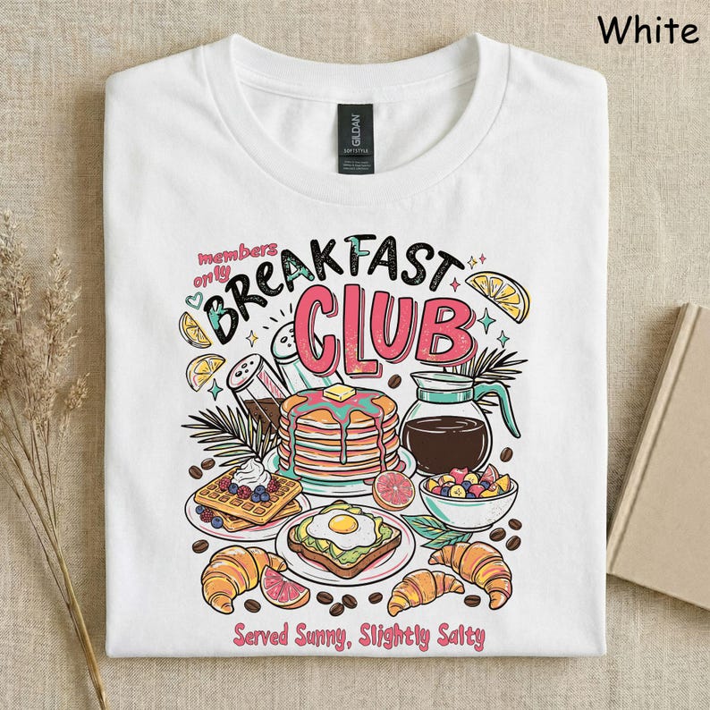 Pode incluir: Camiseta branca com um gr&aacute;fico colorido de alimentos de caf&eacute; da manh&atilde;, incluindo panquecas, waffles e croissants. O texto "Breakfast Club" &eacute; exibido acima das ilustra&ccedil;&otilde;es de comida, com a frase "Served Sunny, Slightly Salty" abaixo.