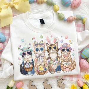 Pode incluir: Camisola branca com um desenho de Páscoa. O desenho mostra quatro gatos com óculos de sol, chapéus e orelhas de coelho, segurando cestas com ovos e um pintinho. As palavras "Happy Easter" estão em um letreiro.