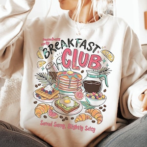 Pode incluir: Camisola creme com um gr&aacute;fico colorido de alimentos de pequeno-almo&ccedil;o, incluindo panquecas, waffles e croissants. O texto "Breakfast Club" &eacute; exibido acima das ilustra&ccedil;&otilde;es de comida, com a frase "Served Sunny, Slightly Salty" abaixo.