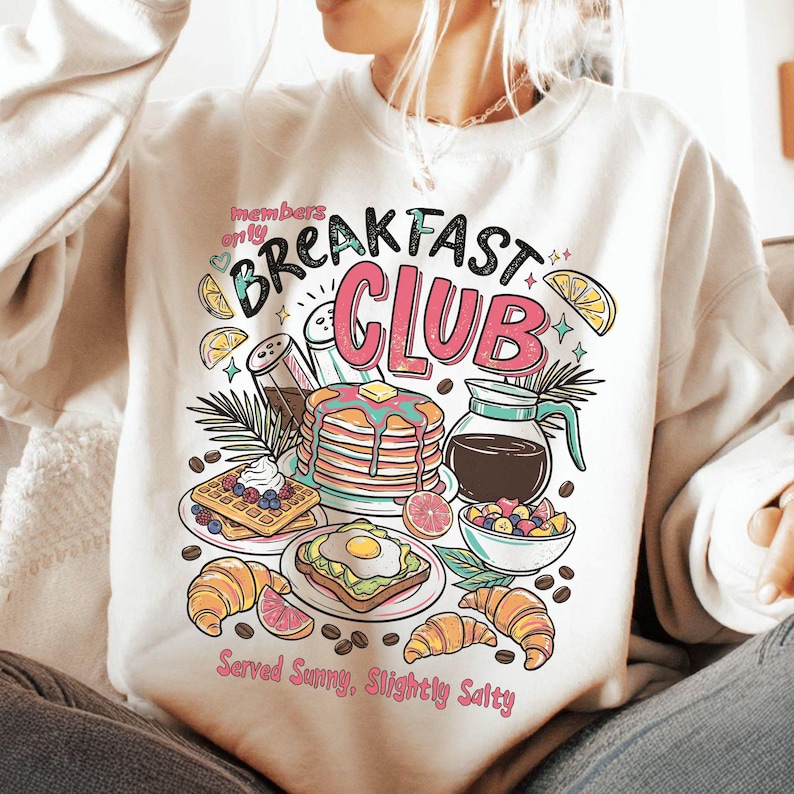 Pode incluir: Camisola creme com um gr&aacute;fico colorido de alimentos de pequeno-almo&ccedil;o, incluindo panquecas, waffles e croissants. O texto "Breakfast Club" &eacute; exibido acima das ilustra&ccedil;&otilde;es de comida, com a frase "Served Sunny, Slightly Salty" abaixo.