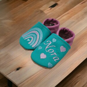 May include: A pair of teal blue baby shoes with a rainbow and hearts embroidered on them. One shoe has the name "Nora" embroidered on it.