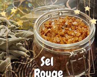 May include: A glass jar filled with reddish-brown coarse salt, labeled "Sel Rouge". The jar has a metal clasp and sits on a patterned surface. Dried sage and small glass containers are in the background, with gold star accents.