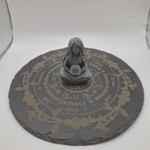 Può includere: Una statuetta grigia di una figura incappucciata che tiene una sfera, seduta su un sottobicchiere rotondo in pietra grigio scuro. Il sottobicchiere presenta un motivo floreale e testo, tra cui le parole "Imbolc" e "Lithia".