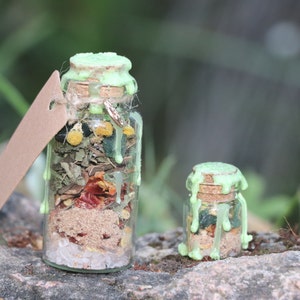 Puede incluir: Dos botellas de vidrio con tapones de corcho y sellos de cera verde. Las botellas est&aacute;n llenas de varias hierbas, especias y cristales. La botella m&aacute;s grande tiene una etiqueta de papel marr&oacute;n adjunta.