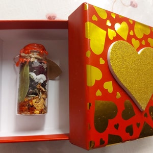 Puede incluir: Una pequeña botella de vidrio llena de hierbas y flores secas, sellada con cera roja, se muestra en una caja roja con un corazón dorado en la tapa. La caja está decorada con corazones dorados sobre un fondo rojo.