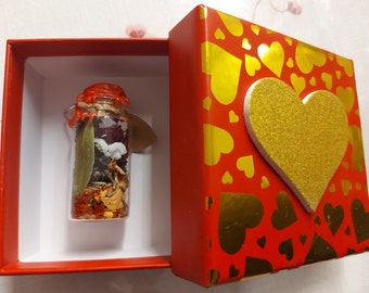 May include: A small glass bottle filled with dried herbs and flowers, sealed with red wax, is displayed in a red box with a gold heart on the lid. The box is decorated with gold hearts on a red background.