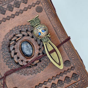 Puede incluir: Un diario de cuero marrón con diseños en relieve intrincados y un marcapáginas decorativo. El marcapáginas presenta un diseño de búho con un emblema del árbol de la vida y una piedra azul. El diario está atado con un cordón granate.