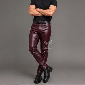 Op de afbeelding: Bordeauxrode leren broek met een slanke pasvorm, een zwarte riem met een zilveren gesp en zichtbare stiksels. De broek wordt gecombineerd met een zwart t-shirt en zwarte laarzen. Een logo met de tekst LEATHER CRAFTERY is zichtbaar op het been.