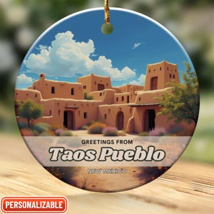 May include: A round ceramic ornament with a colorful image of Taos Pueblo, New Mexico. The ornament features a blue sky with white clouds, a brown adobe building, and green trees. The text "Greetings from Taos Pueblo, New Mexico" is printed on the ornament.