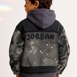 May include: A denim jacket with a gray tie-dye pattern and the name "JORDAN" printed on the back. The jacket has a gray hood and is worn by a child. The jacket has a distressed, splattered design.