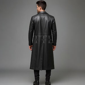May include: A full-length black leather coat with a notched collar. The coat has a tailored fit and a long, flowing design. The back of the coat features a vent at the hem. The coat is worn with black pants and shoes. The Tannery Lane logo is visible.